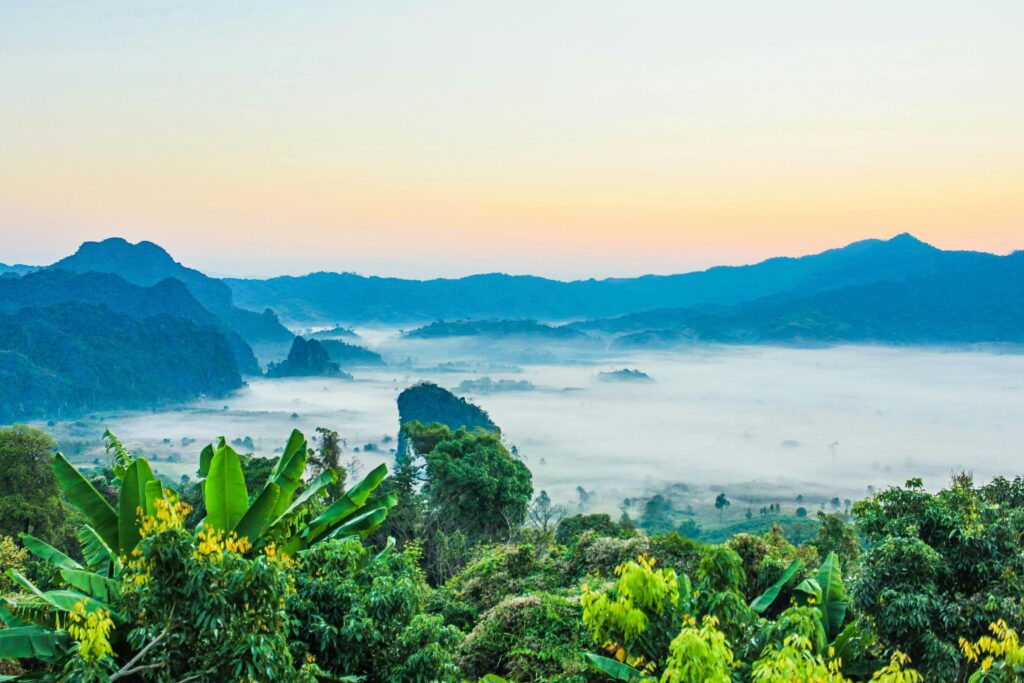 Isaan Motorcycle Tour: A Journey into Thailand’s Heartland Scenic view of misty valleys surrounded by green hills and banana trees under a pastel sunrise sky