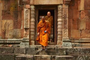 Two Buddhist monks wearing orange robes stepping out of an ancient stone temple with intricate carvings